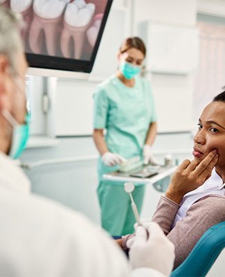 Woman in teal dental chair holding two fingers to her cheek looking at dentist in white coat
