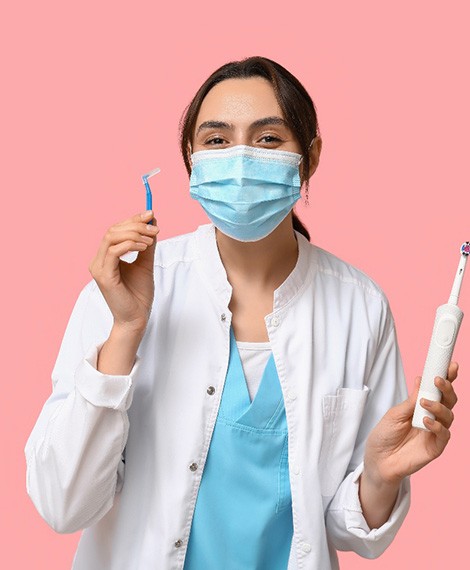 Dentist in facemask holding electric toothbrush in one hand and interproximal brush in the other